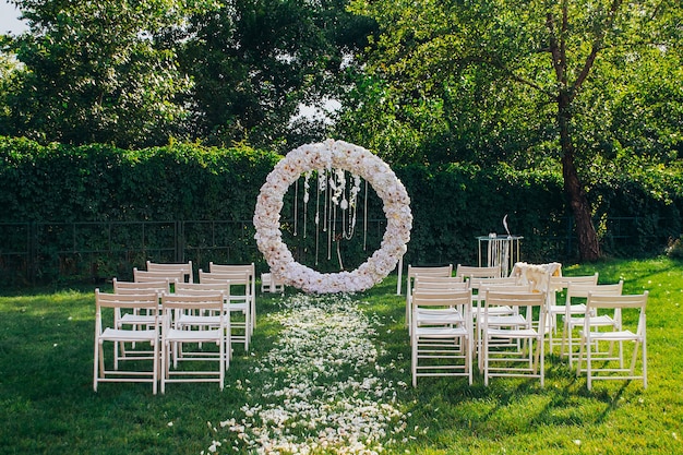 Foto arco nuziale rotondo fatto di fiori bianchi con perline e piume su uno sfondo di verde accanto a sedie di legno bianco