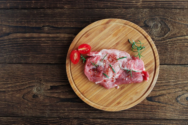 round wooden cutting board lies a raw pickled pork steak. Near a sprig of rosemary and cherry tomatoes.