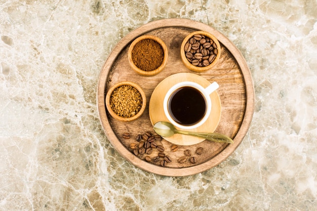 Round wooden tray with a cup of dark coffee and with various kinds of coffee in mini bowls. top view. marble background.