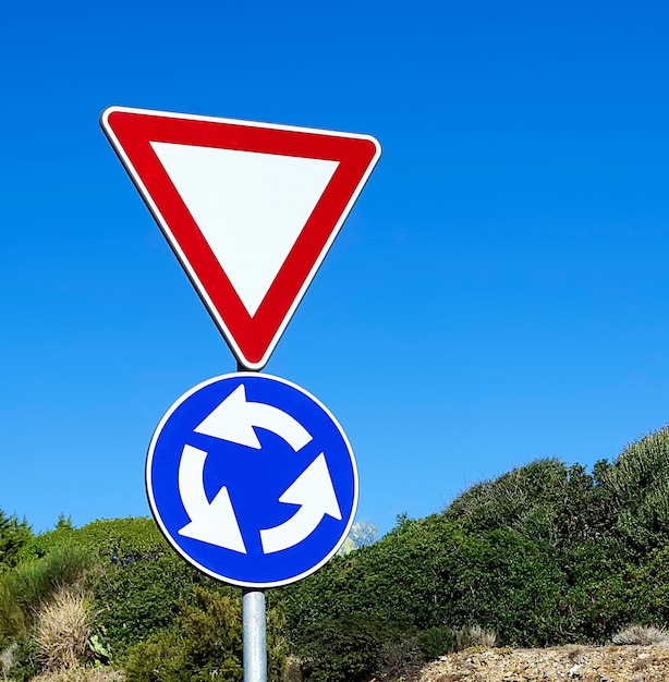 Roundabout and give away signs surrounded by vegetation under a blue sky