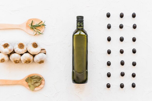 Row of black olives with oil bottle, garlic bulbs, and herbs on white textured backdrop