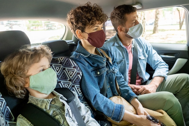 Row of contemporary young family of three in protective masks and denim jackets looking forwards while going by car on backseat