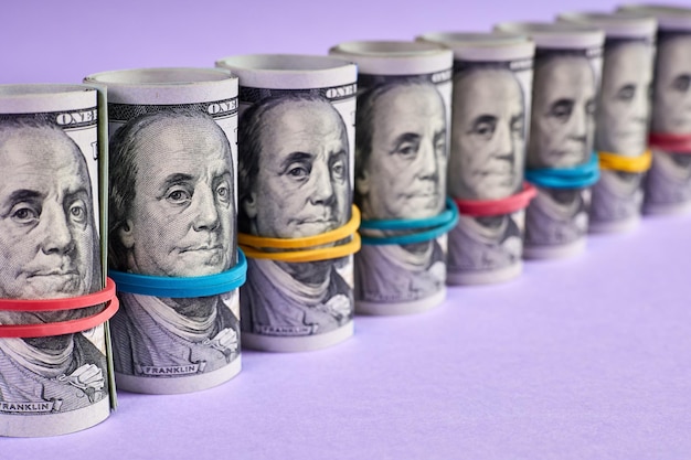 A row of rolled hundred dollar bills with rubber bands on purple background