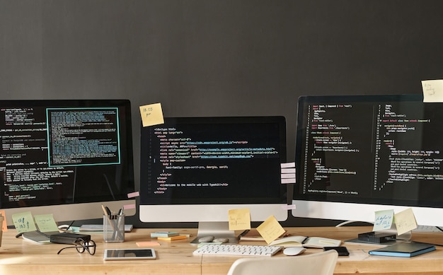 Row of three computer monitors with coded data and notepapers on their screens standing on table in