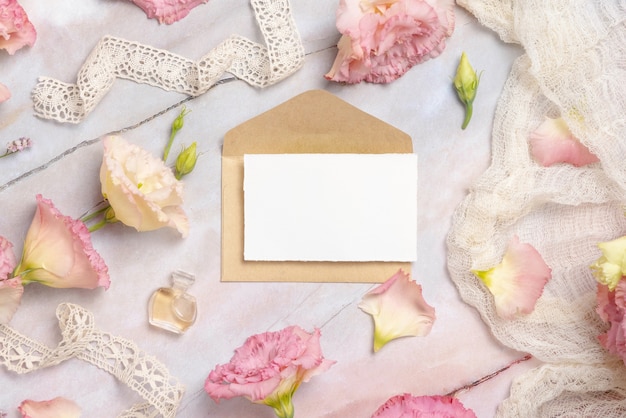 Roze bloemen en een blanco wenskaart met envelop op een marmeren tafel versierd met vintage linten, bloemblaadjes en parfumflacon. Mock-up scène. Vrouwelijk plat gelegd