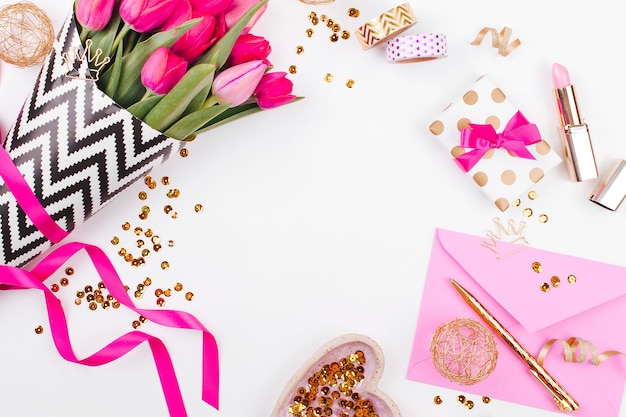 Roze en goud gestyled bureau met bloemen. Roze tulpen in zwart-wit stijlvol inpakpapier, geschenken, cosmetica en vrouwelijke accessoires met confetti op witte achtergrond