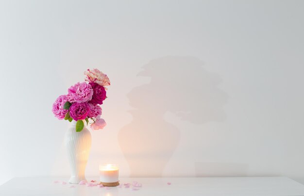Roze rozen in vaas en brandende kaars op witte muur als achtergrond
