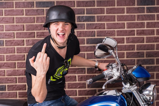 Foto giovane aggressivo maleducato che si siede sulla sua moto nel casco che fa un gesto offensivo scortese con il dito medio mentre grida alla telecamera