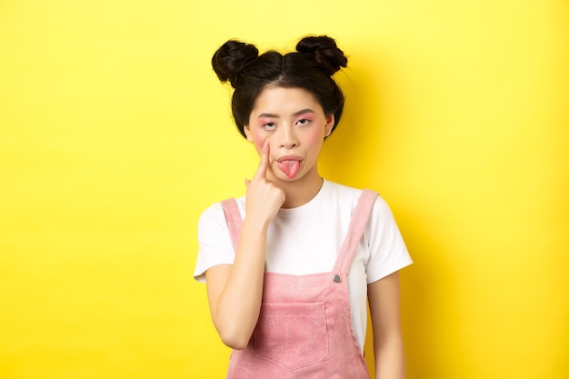 Rude asian girl stretch eyelid and showing tongue, mocking someone, standing on yellow.