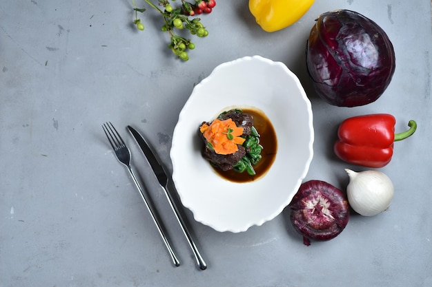 Rundvlees wangen. Op een grijze tafel staat een bord met een schotel vlees in een donkere saus met spinazie en worteltjes