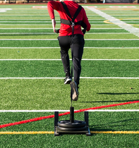 Runner is pulling a weighted sled across a green turf field for speed and strength training