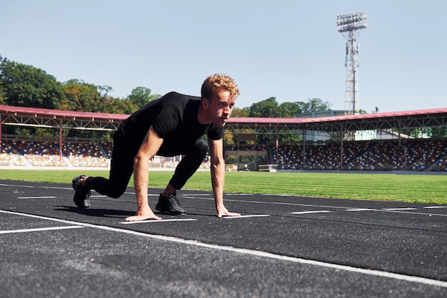 Runner is on starting position on track Sportive young guy in black shirt and pants outdoors at daytime