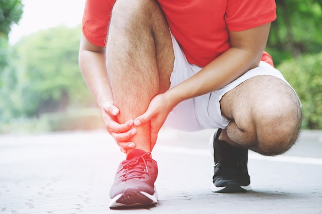 Бегун трогает болезненно скрученную или сломанную лодыжку. Несчастный случай на тренировке спортсмена-бегуна. Спортивный бег растяжение связок лодыжки вызвало травму колена. и боль в костях ног. Сосредоточьте внимание на красных ногах, чтобы показать боль.