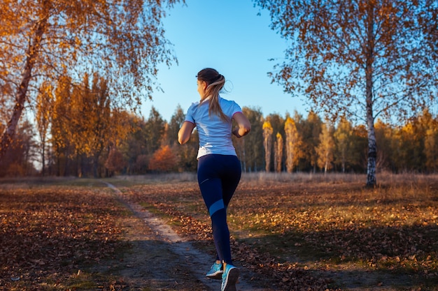 秋の公園でランナートレーニング。日没時の水のボトルで走っている女性。アクティブなライフスタイル