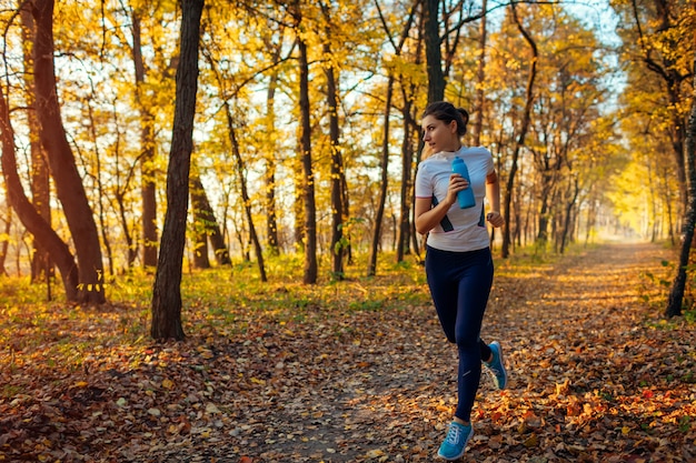 Runner uitoefenen in herfst park. Vrouw die met waterfles bij zonsondergang loopt. Actieve gezonde levensstijl