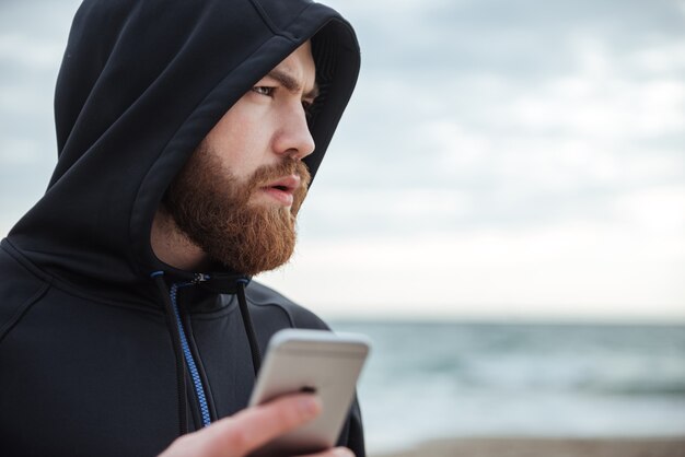 Runner with phone on beach in profile man in hood