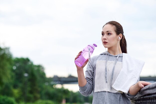 Running asian woman is having break drinking water during run in summer park