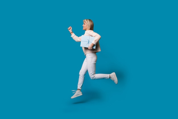 Running blonde woman is holding a present box on a blue studio wall and smile in casual clothes