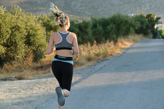 Esecuzione di ragazza fitness in cuffie con smartphone, vista posteriore, copia dello spazio. soleggiata giornata estiva, strada in montagna, stile di vita sano attivo nei giovani