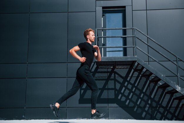 Photo running near black building sportive young guy in black shirt and pants outdoors at daytime