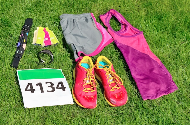 Running shoes, marathon race bib (number), runner gear and energy gels on grass, sport competition, fitness and healthy lifestyle concept