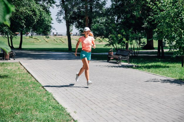 Running smiling young sporty woman in park in sportswear in front of green trees on road of grey til...