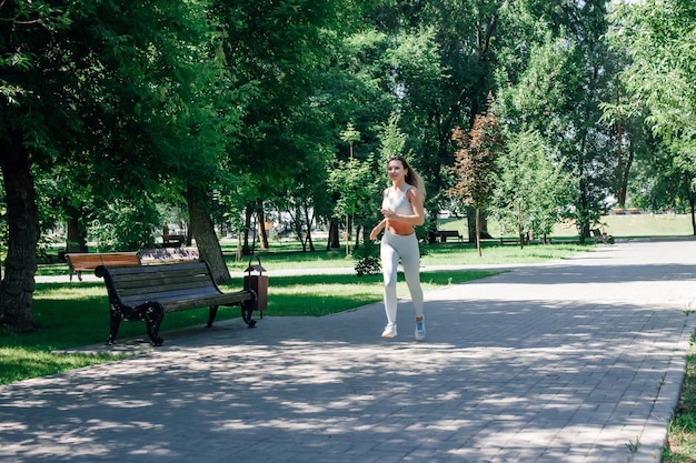 Running smiling young sporty woman warming up stretching in grey sportswear in park in sunny summer day