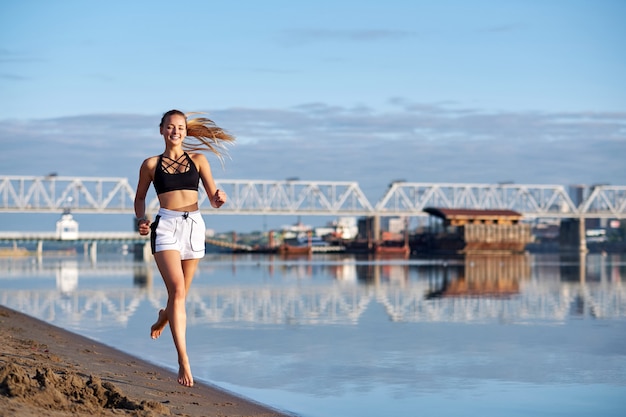 日の出の砂の中を走っている女性。都市の背景にビーチや川の海岸で裸足でジョギングする朝