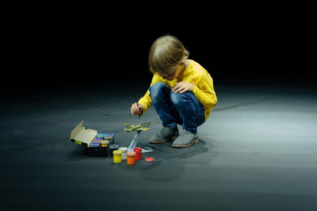 Russia Moscow 06152022 candid little six year old kid boy painting or colorize or colour model airplane with gouache paints while squatting on floor studio illuminated by white yellow bright light