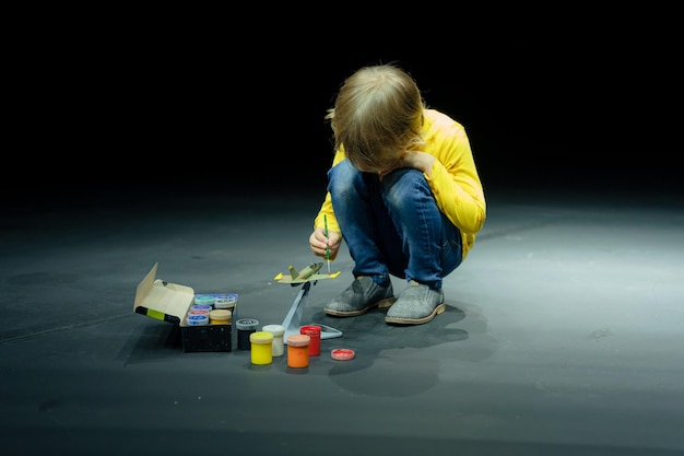 Russia Moscow 06152022 candid little six year old kid boy painting or colorize or colour model airplane with gouache paints while squatting on floor studio illuminated by white yellow bright light