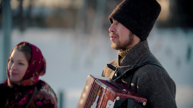 ロシアの民間伝承ロシアのひげを生やした男性が屋外でアコーディオンを演奏し、女性が彼のそばに立っている