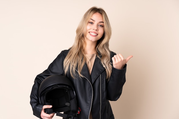 La ragazza russa con un casco del motociclo sulla parete beige con i pollici aumenta il gesto e sorridere