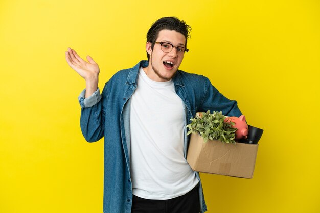 Russian Man making a move while picking up a box full of things isolated on yellow background having doubts while raising hands
