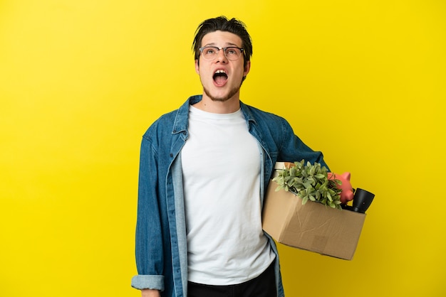 Russian Man making a move while picking up a box full of things isolated on yellow background looking up and with surprised expression