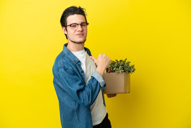 Russian Man making a move while picking up a box full of things isolated on yellow background proud and self-satisfied