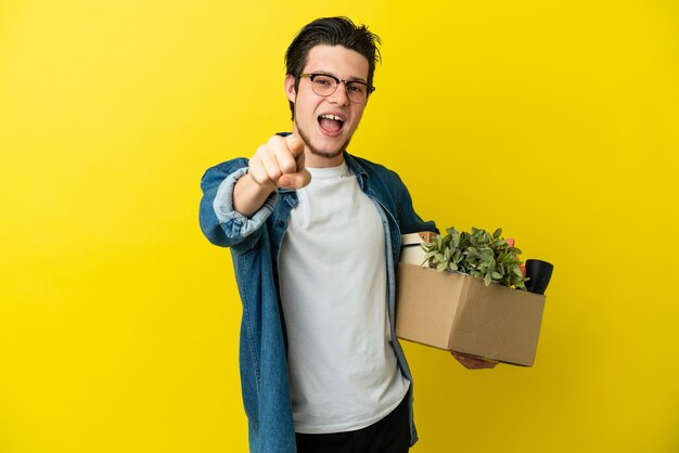 Russian Man making a move while picking up a box full of things isolated on yellow background surprised and pointing front