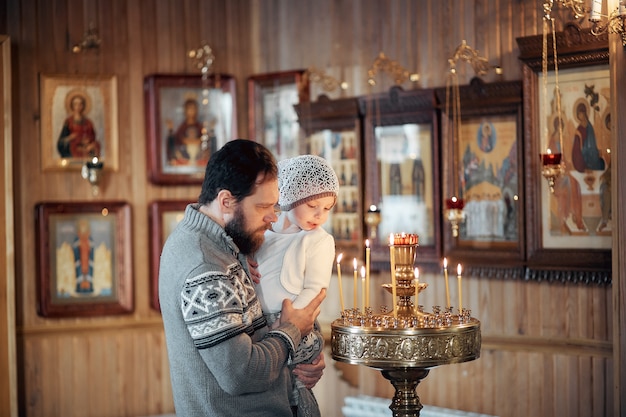 Русский мужчина с бородой и дочерью стоит в православной церкви, зажигает свечу и молится перед иконой.