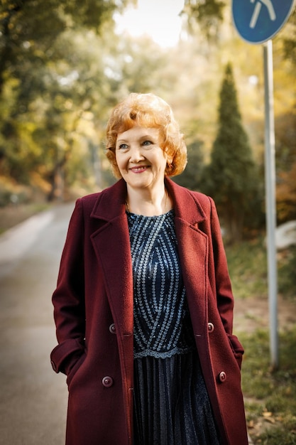 Russian mature woman with short hair smiling looks to the side outdoors in autumn in an unbuttoned burgundy coat