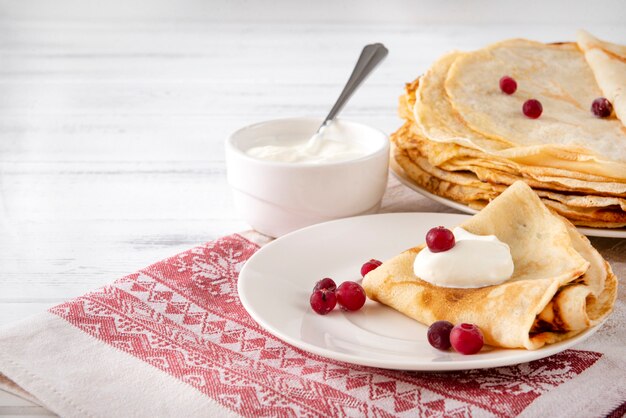 Фото Русские оладьи с ягодами и сметаной на тарелке, на полотенце с красным рисунком на белой поверхности,