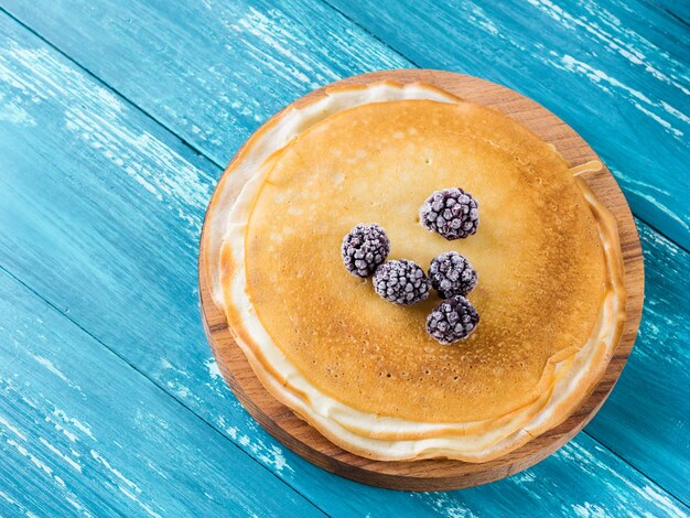 Photo russian pancakes on a wooden plate on an azure table with frozen blackberries.