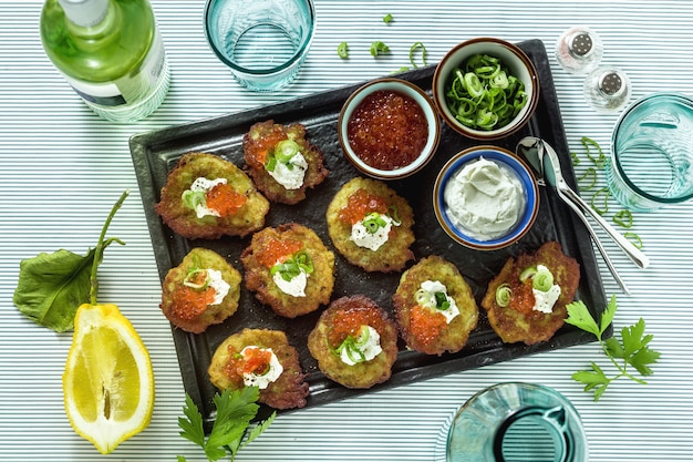 Russian traditional potato pancakes served with red caviar, green onions and sour cream on a tray with white wine and lemon