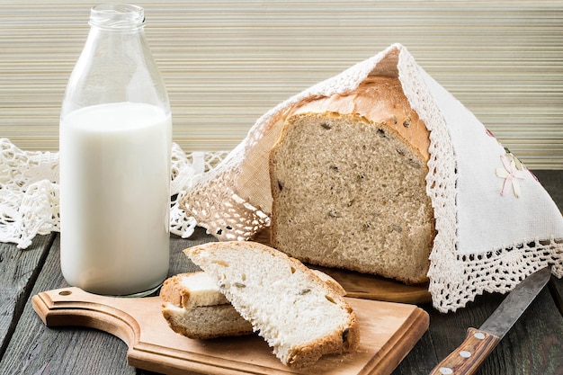 Russian wholegrain homemade wheat bread with sunflower seeds on the board under the cloth with knitted border, milk in a bottle on wooden table