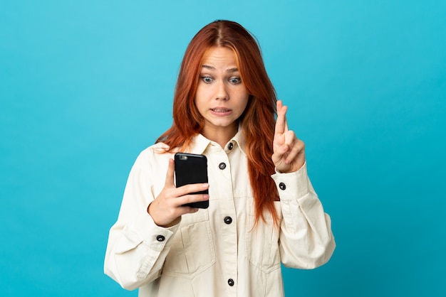 Русская женщина изолирована на синем, используя мобильный телефон со скрещенными пальцами