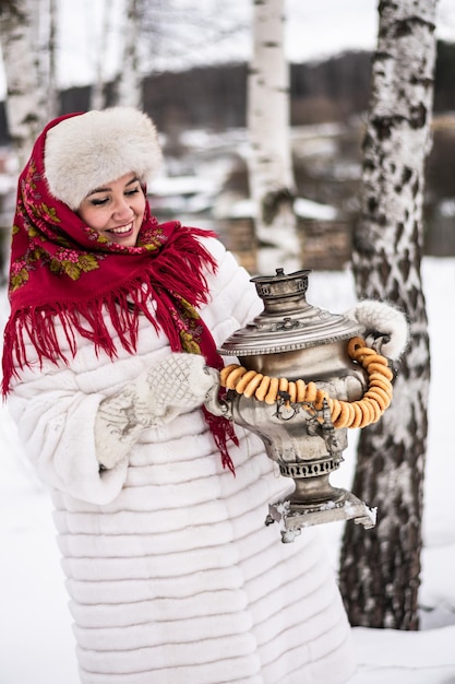 Russian woman in a shawl and a white fur coat with a samovar in\
a field among birches