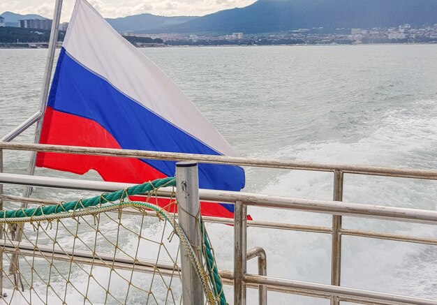 Russische driekleurige vlag op een schip, tegen de achtergrond van de zee en de kustlijn met een kuststad, dageraad, zomer.