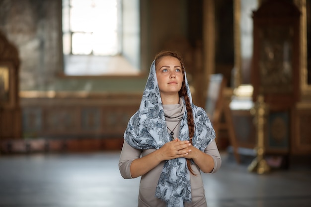 Russische mooie blanke vrouw met rood haar en een sjaal op haar hoofd is in de orthodoxe kerk, steekt een kaars aan en bidt voor het pictogram.