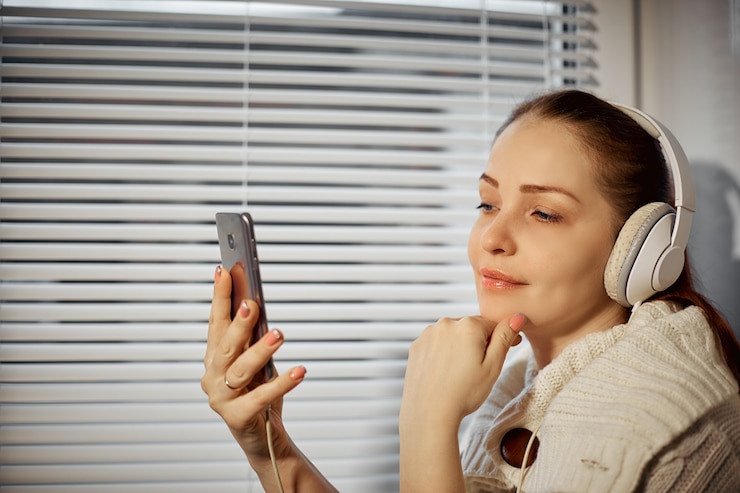 Rust en ontspanning, jonge blanke vrouw luisteren naar muziek in koptelefoon met behulp van een smartphone zittend op de vensterbank.
