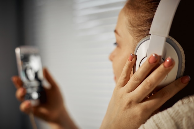 Foto rust en ontspanning, jonge blanke vrouw luisteren naar muziek in koptelefoon met behulp van een smartphone zittend op de vensterbank.