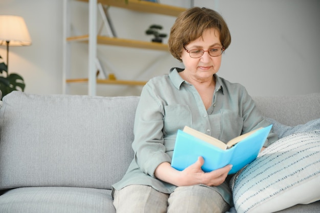 Rustgevend lezen Portret van een doordachte oude vrouw die favoriete literatuur leest in een gezellig huis Ze ligt op kussens op een comfortabele bank Benen zijn bedekt met een zachte deken