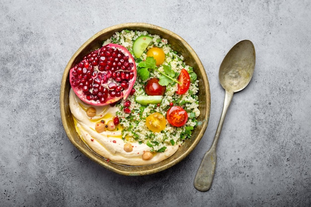 Rustic bowl with couscous salad with vegetables, hummus and fresh cut pomegranate. Middle eastern or Arab style meal with seasonings and fresh cilantro. Beautiful and healthy Mediterranean dinner